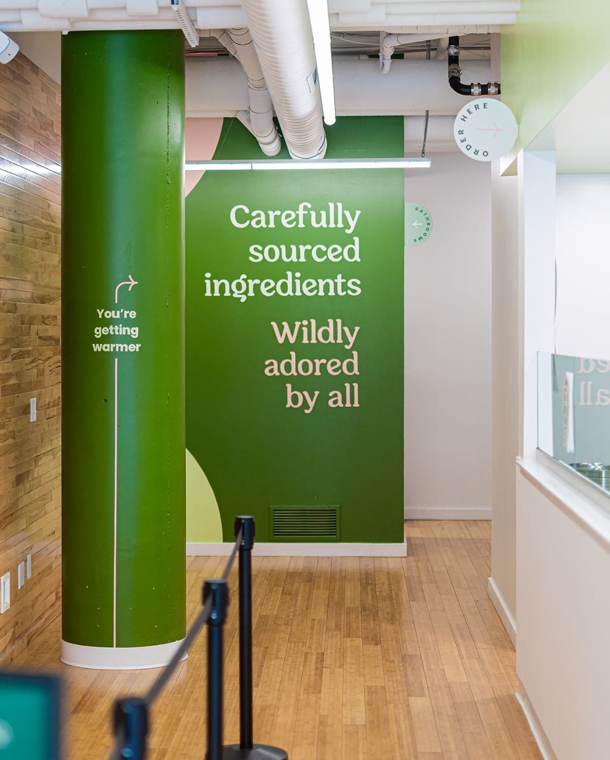 Interior of a modern restaurant with green walls and wood flooring. A large green pillar has the text "You're getting warmer" with an arrow. On the back wall, text reads "Carefully sourced ingredients" and "Wildly adored by all" in white and light pink font. Ceiling pipes and light fixtures are visible, and signs indicate directions to order and bathrooms.