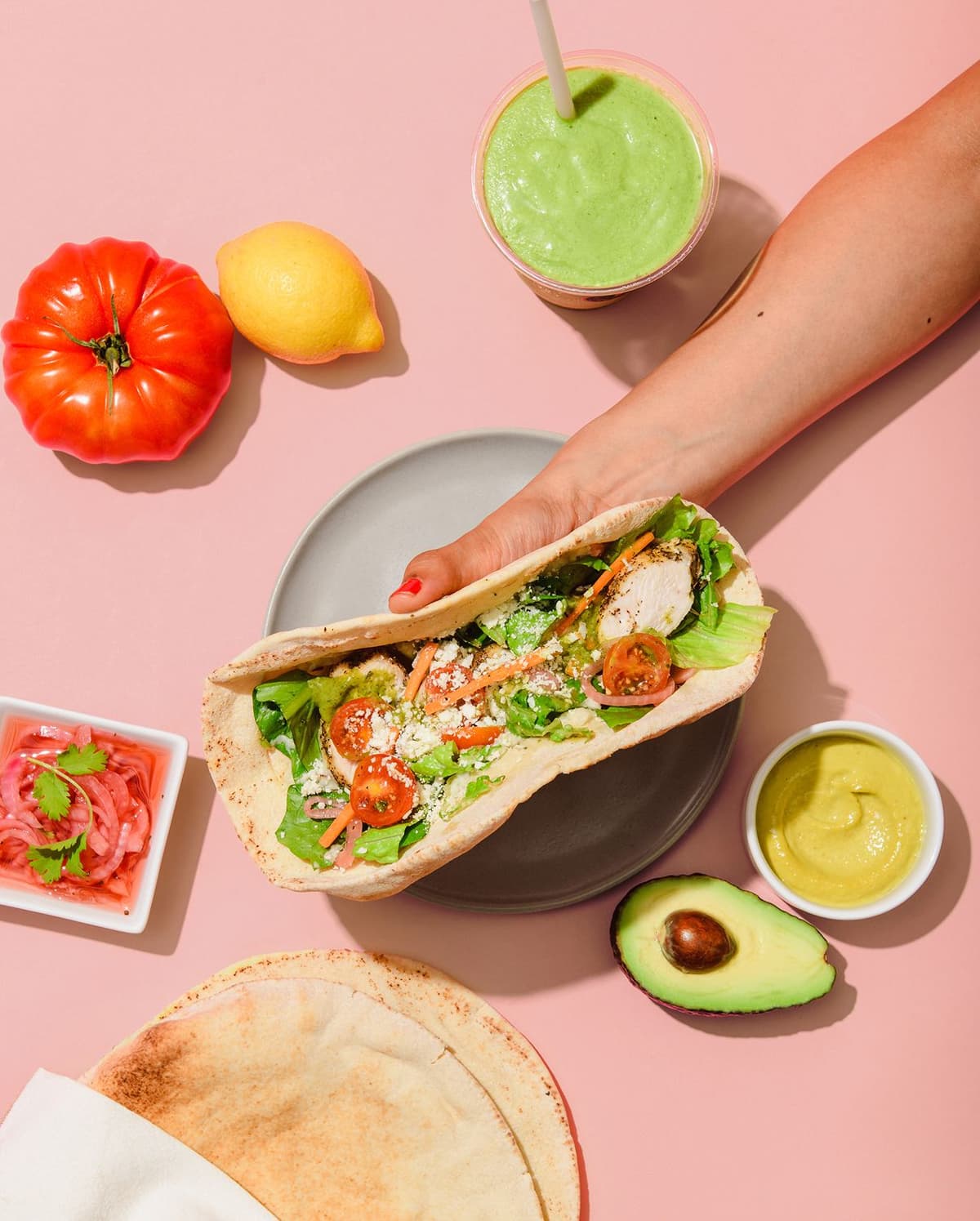 Hand holding a pita wrap filled with grilled chicken, lettuce, cherry tomatoes, shredded carrots, and cheese, placed over a grey plate on a pink surface. Surrounding are a red tomato, a lemon, a cup of green smoothie with a straw, a small white bowl with mustard sauce, half an avocado, a small white square bowl with pickled onions garnished with cilantro, and two stacked pita breads partially wrapped in a napkin.