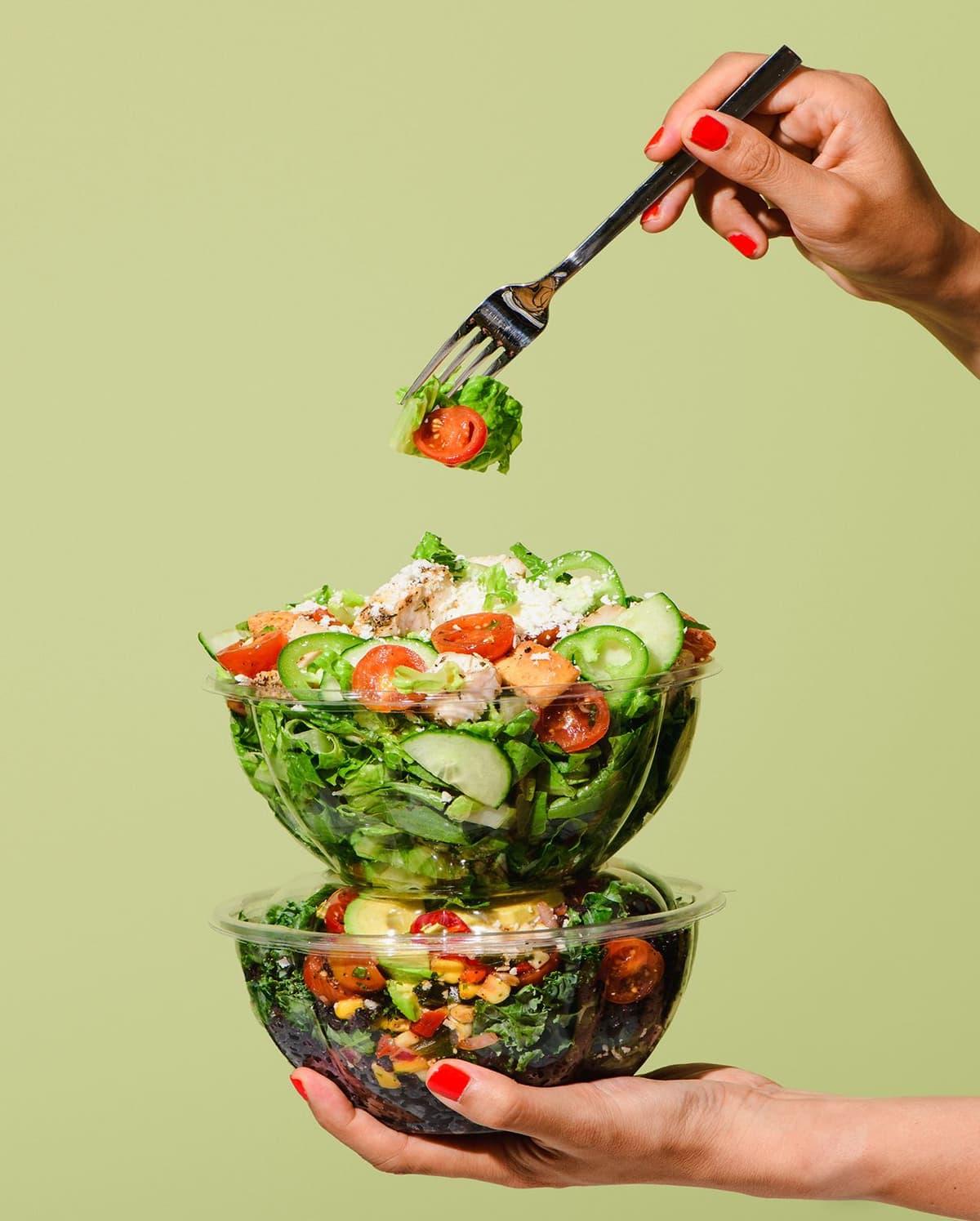 Hand with red nail polish holding a fork with a bite of salad above two stacked clear plastic bowls filled with fresh salad containing lettuce, cherry tomatoes, cucumber, grilled chicken, and cheese, against a light green background.