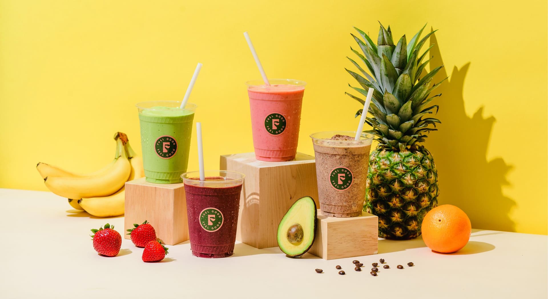 Four colorful smoothies in clear plastic cups with straws, each placed on or near wooden blocks, accompanied by fresh fruits including bananas, strawberries, avocado, pineapple, and an orange, set against a bright yellow background.