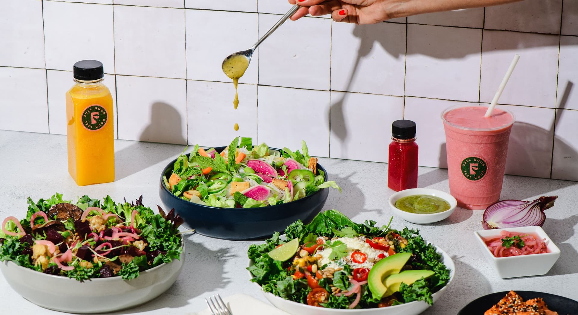 A tabletop with three bowls of fresh salads containing leafy greens, avocado, cherry tomatoes, watermelon radish, and red chili slices. A hand is drizzling dressing from a spoon over the middle salad. On the left, there is a bottle of orange juice labeled "Fuel Good". On the right, there is a pink smoothie with a straw, a small red shot bottle, a small dish of green sauce, a small bowl of pickled onions, and a halved red onion. Some crackers are in the bottom right corner. The background is a tiled white wall.