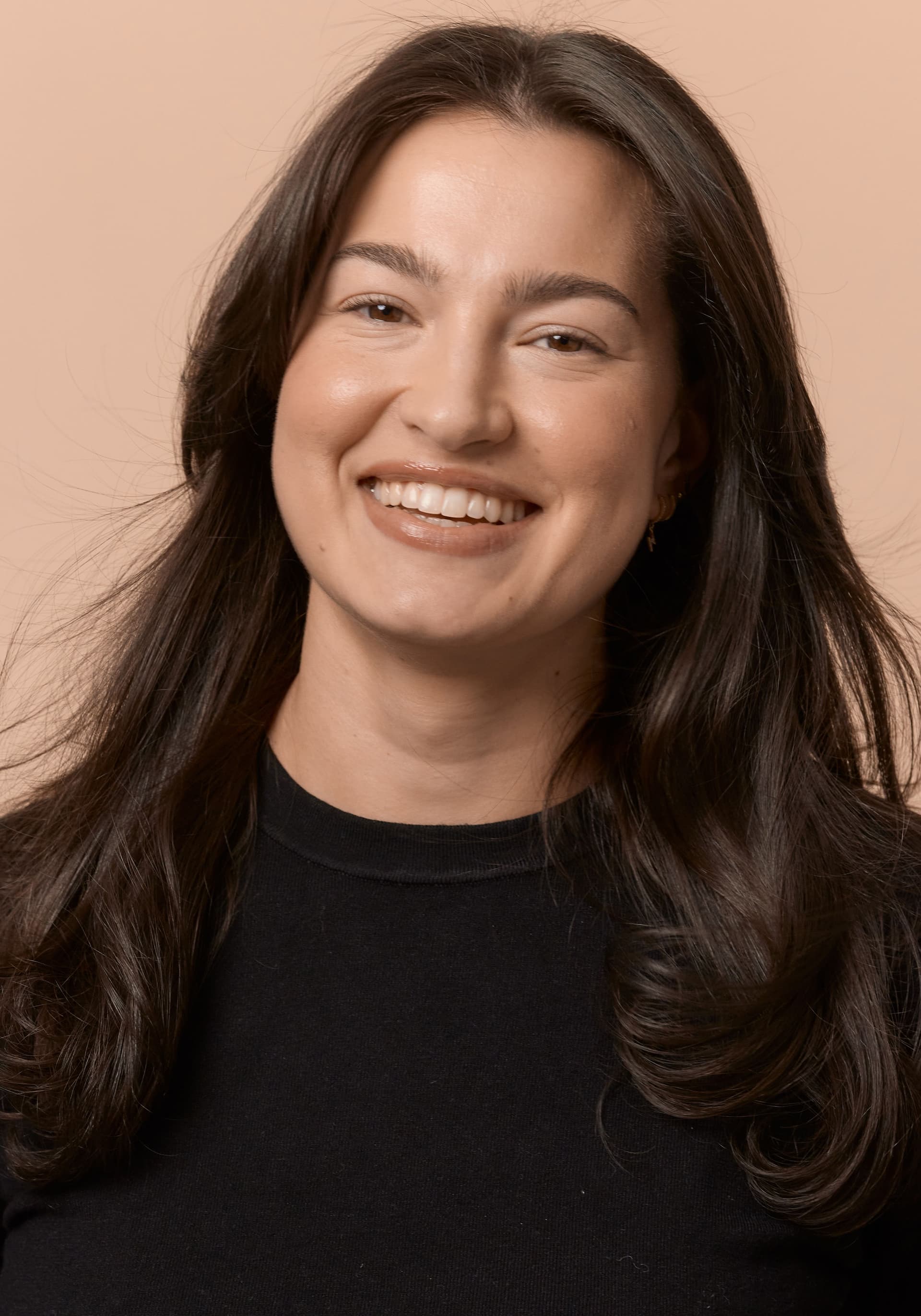 Portrait of a young woman with long dark brown hair, smiling, wearing a black top against a beige background.