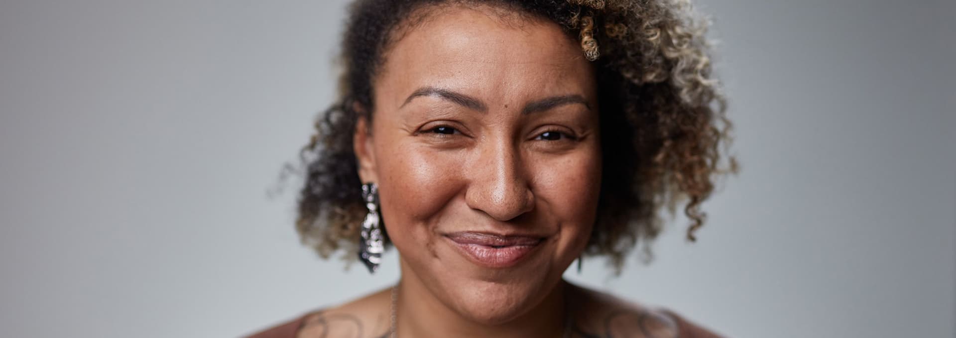 Close-up portrait of a woman with curly hair, wearing dangling earrings and a subtle smile against a plain gray background.
