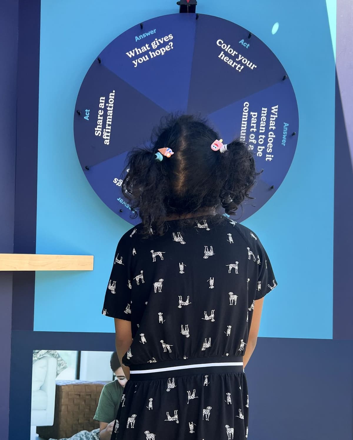 A young girl with curly hair in pigtails and wearing a black dress with white dog prints stands facing a blue wall-mounted spinning wheel with questions and actions written on it like "What gives you hope?" and "Color your heart!"