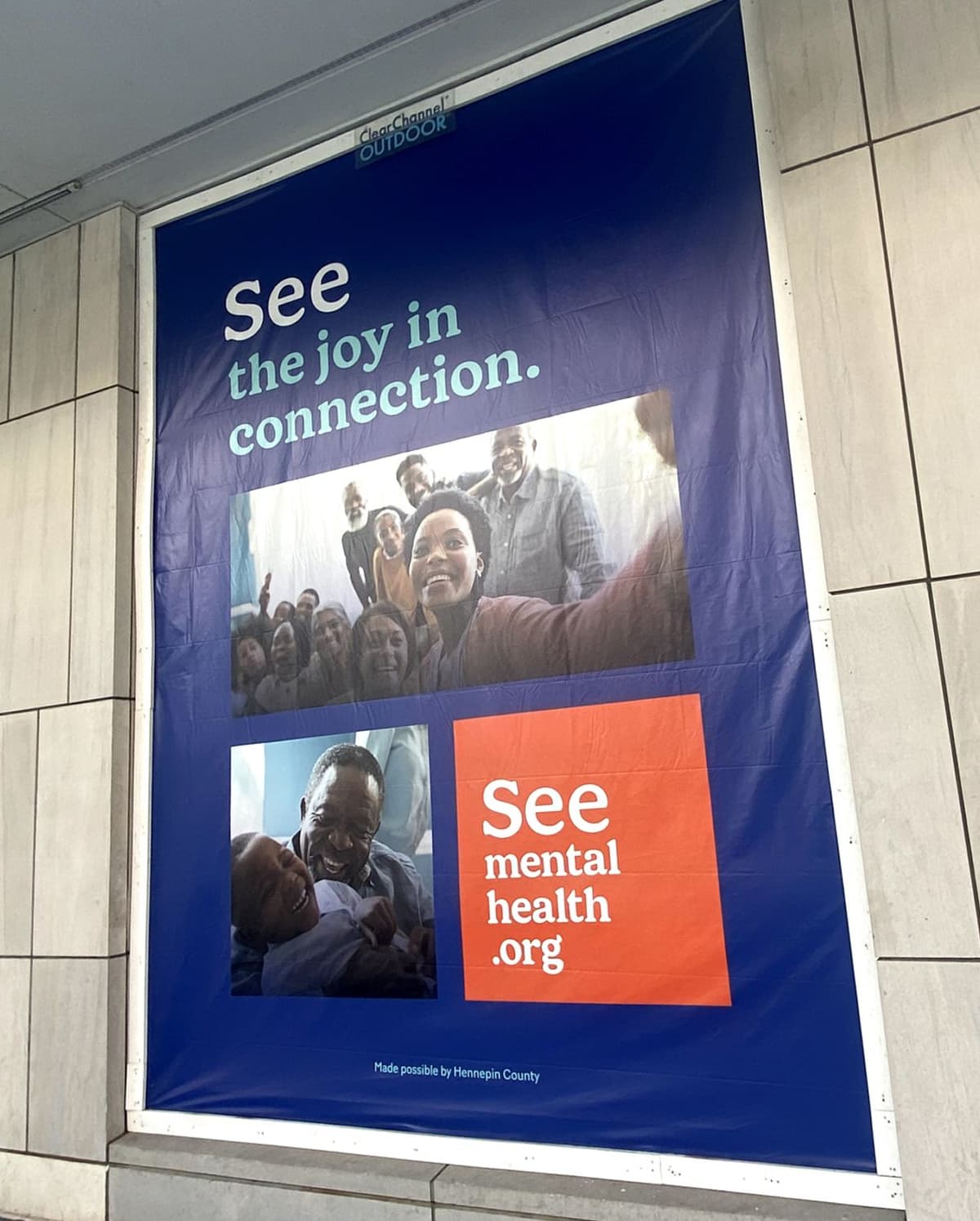 Poster on a tiled wall with a blue background and text that reads "See the joy in connection." It includes two photos: one of a large group of people smiling and taking a selfie, and one of an older man playfully interacting with a young child. The bottom right features an orange box with the text "See mental health .org." The poster is credited to Hennepin County.