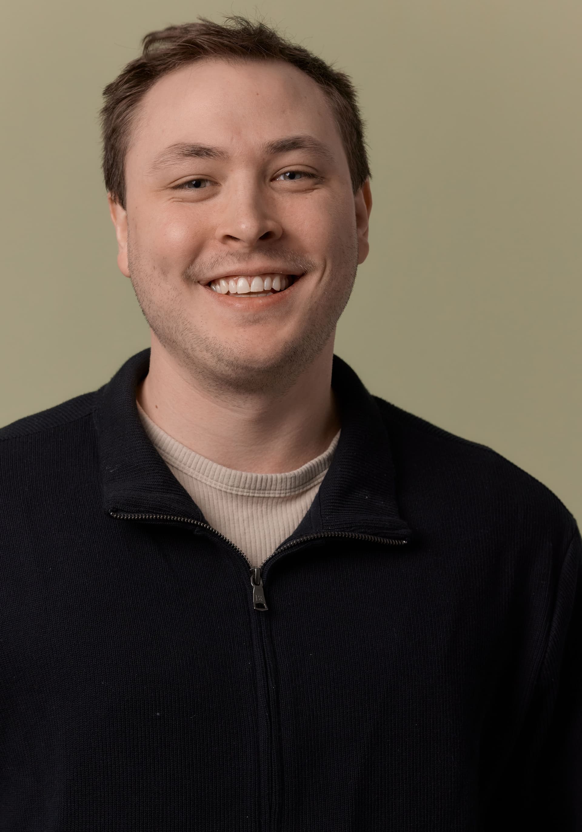 Smiling young man with short brown hair wearing a black zip-up sweater over a beige ribbed shirt, against a plain beige background.