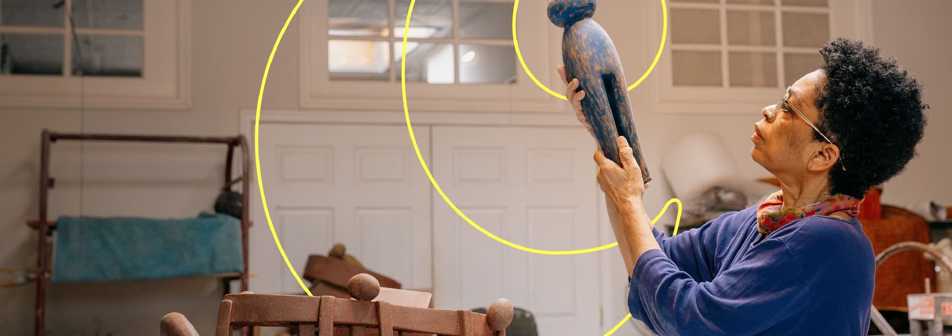 Person with short curly hair and glasses holding and examining a blue wooden humanoid sculpture in a room with white paneled doors and various pieces of furniture.