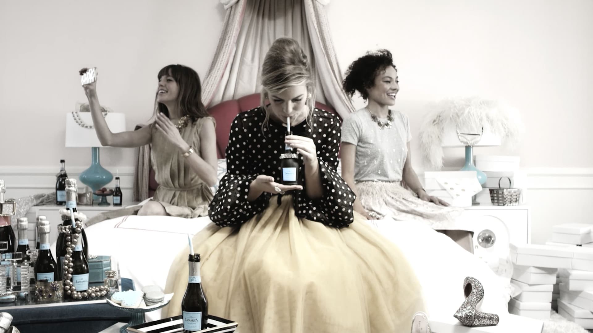 Three women sitting on a bed in an elegant, softly lit room. The woman in the center, wearing a black polka-dot top and yellow skirt, is drinking from a bottle with a straw. The woman on the left, in a beige dress, is holding up a phone, seemingly taking a selfie. The woman on the right, in a light-colored dress, is smiling and looking to the side. Bottles, jewelry, and gift boxes are scattered around the room, suggesting a celebratory or party atmosphere.