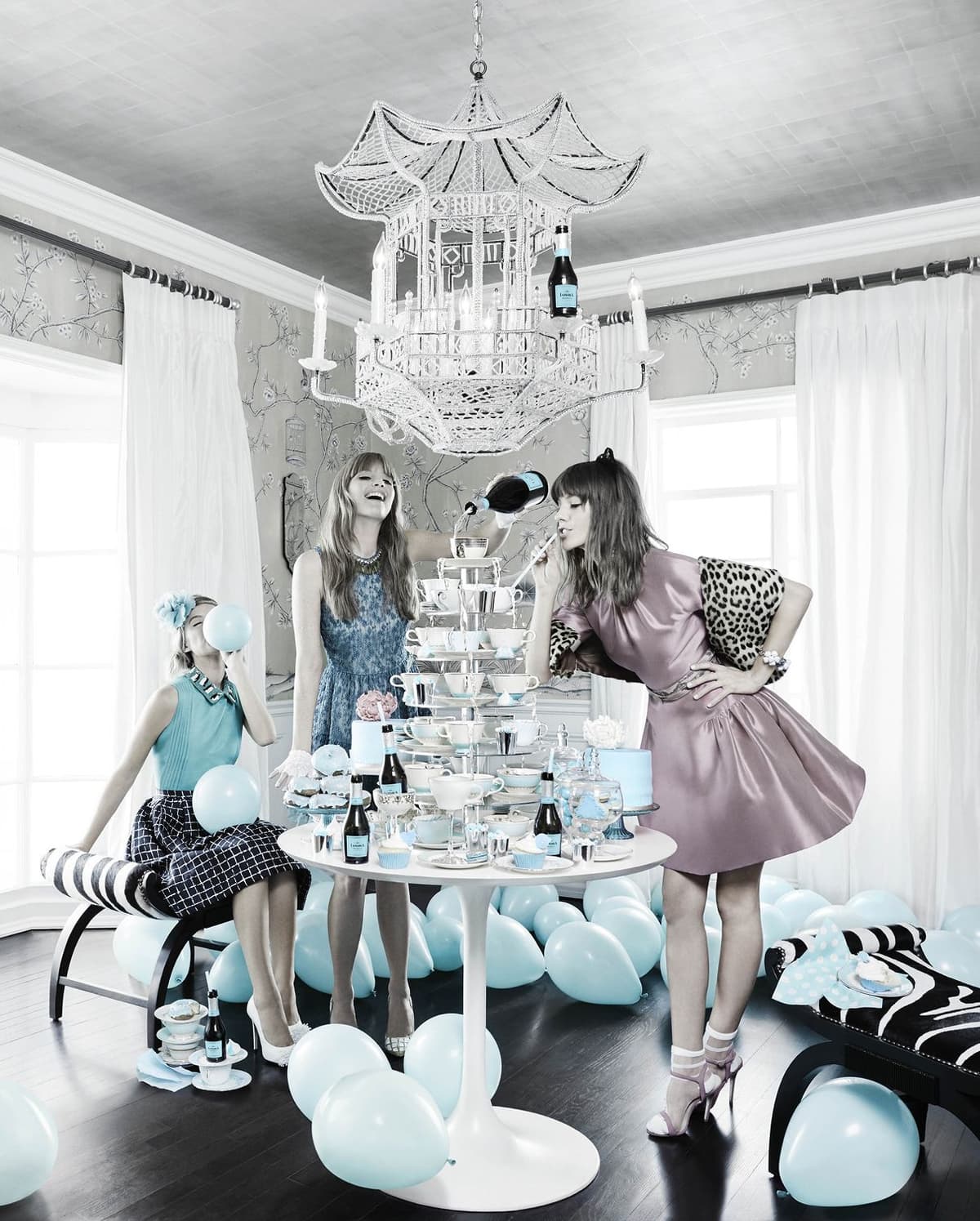 Three women in a decorated room with light blue balloons scattered on the floor. One woman sits on a striped bench blowing a blue balloon, wearing a turquoise top and patterned skirt. A second woman in a blue dress is pouring a drink into a tall stack of teacups on a round white table. The third woman, dressed in a pink satin dress with a leopard print sleeve, leans in to sip with a straw from the teacup stack. A large white chandelier hangs overhead and the room has floral wallpaper and white curtains.