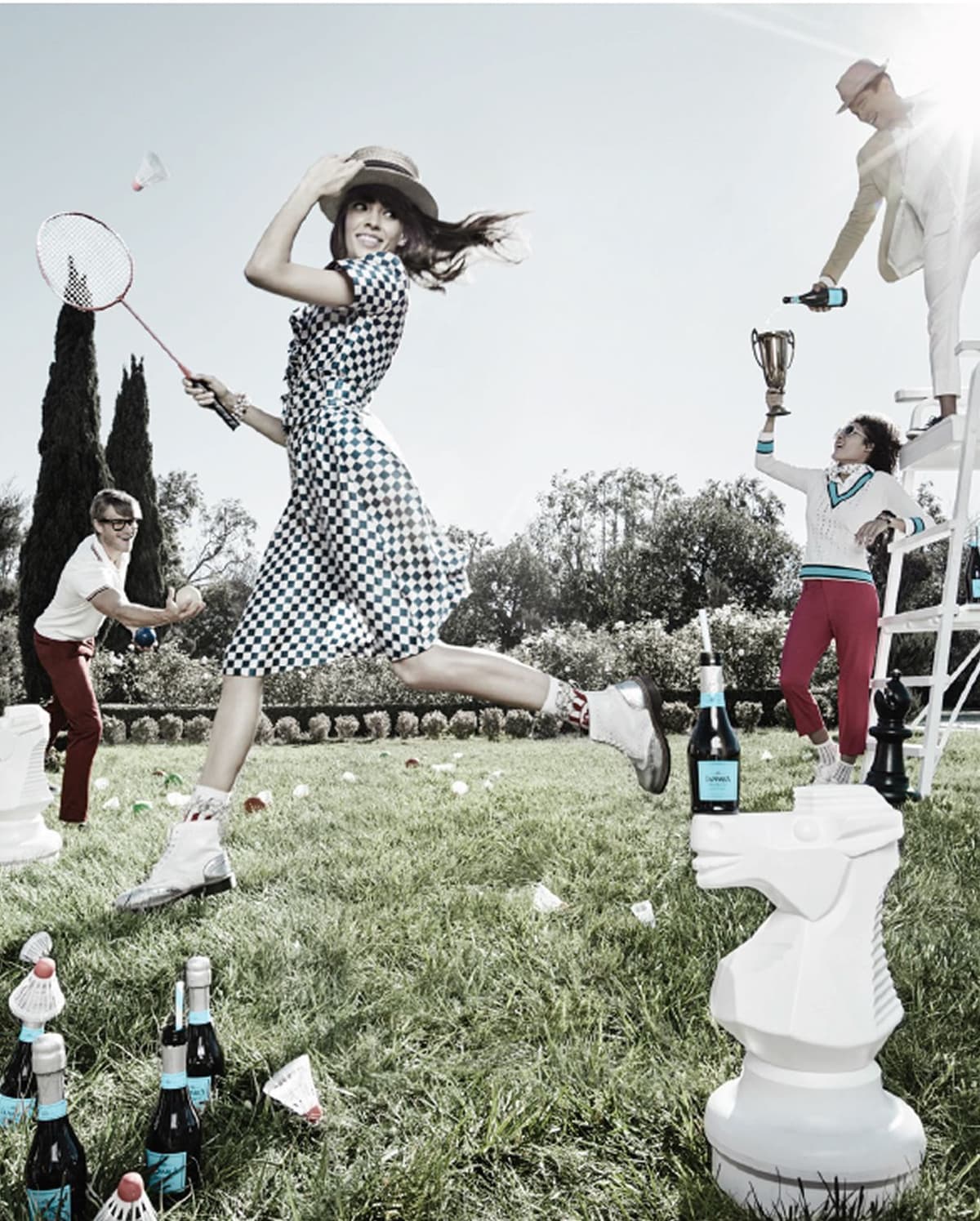 A woman in a checkered dress and hat playing badminton on grass with a man preparing to hit a ball nearby. Another person stands on an elevated chair holding a bottle, while a person on the ground raises a trophy. Large chess pieces and bottles are scattered around the scene, set outdoors with trees in the background.