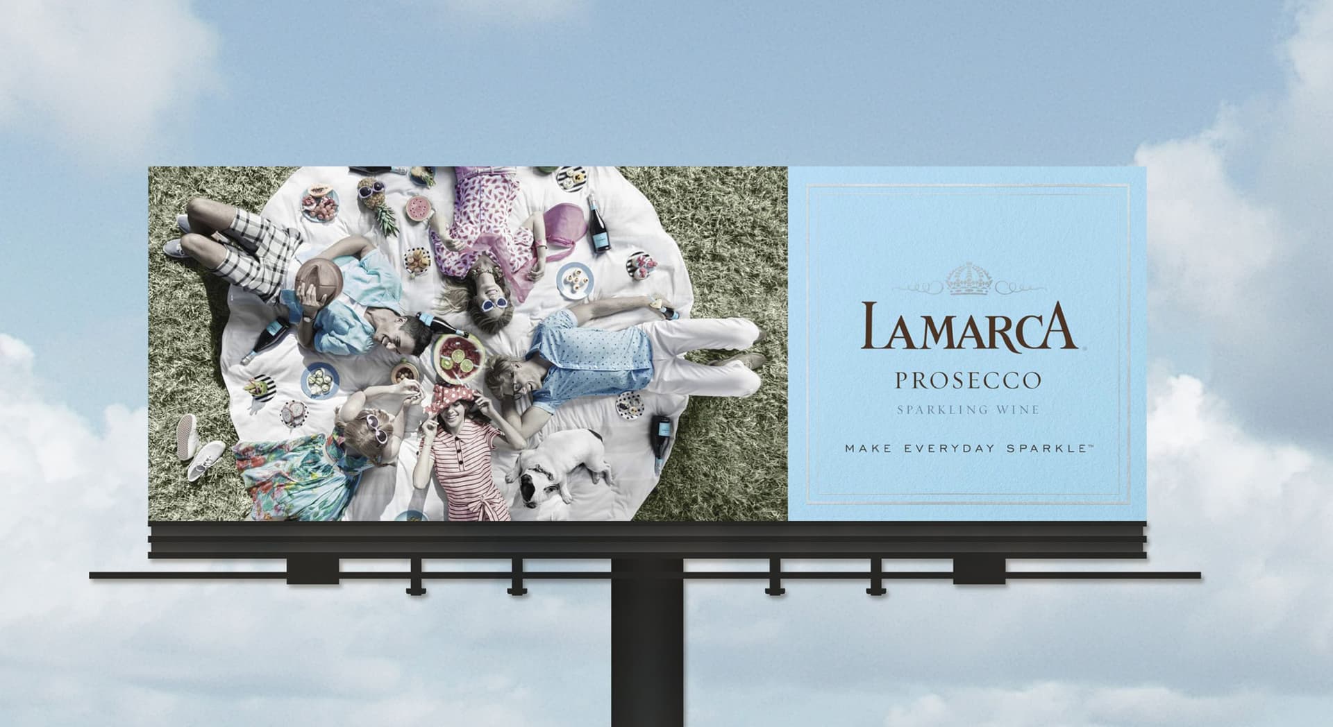 Billboard featuring an aerial view of five people lying on a white blanket on grass, surrounded by picnic food and bottles of La Marca Prosecco, with a dog resting nearby. The right side of the billboard has a blue background with the La Marca Prosecco logo and the slogan "MAKE EVERYDAY SPARKLE™."
