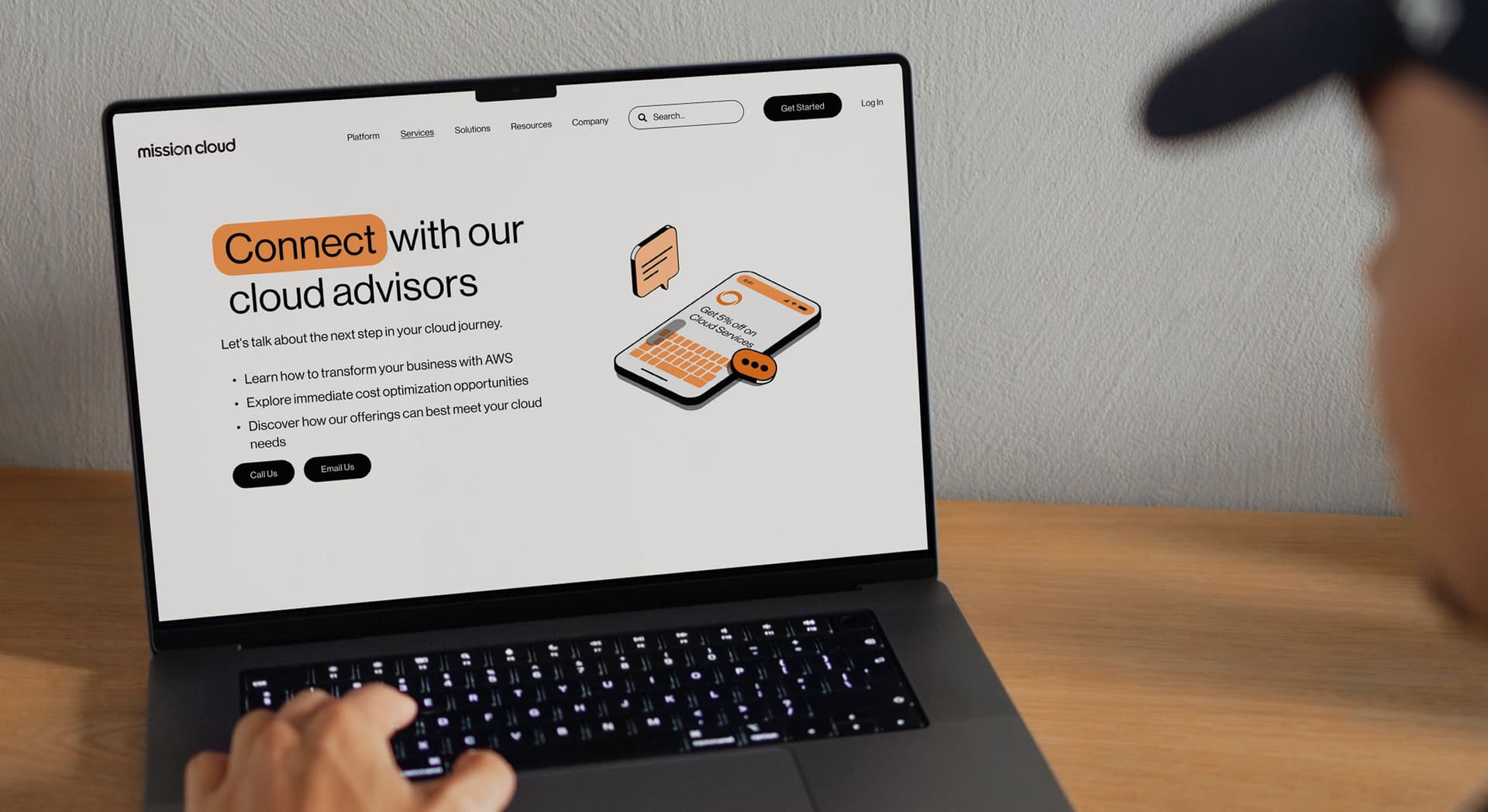 Person wearing a dark cap is using a laptop displaying a webpage for "mission cloud" with the heading "Connect with our cloud advisors" and options to call or email for cloud services.