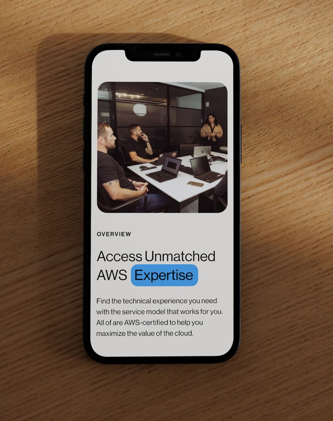 Smartphone on a wooden surface displaying a webpage with a photo of four people in a modern office meeting room, working on laptops. The webpage headline reads "Access Unmatched AWS Expertise" with a highlighted blue background behind the word "Expertise." The text below promotes AWS-certified technical experience and cloud service models.