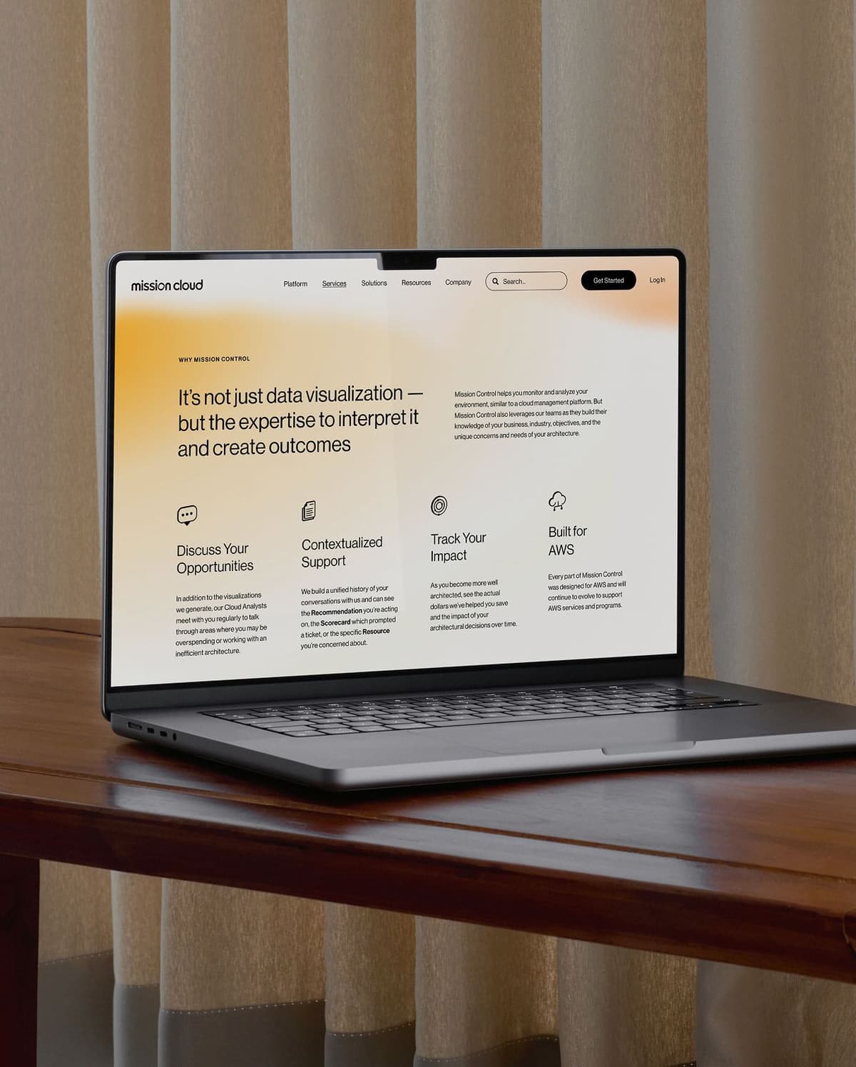 Laptop on wooden desk displaying a website called "mission cloud" with a headline about data visualization expertise and sections titled Discuss Your Opportunities, Contextualized Support, Track Your Impact, and Built for AWS. Beige curtains are in the background.