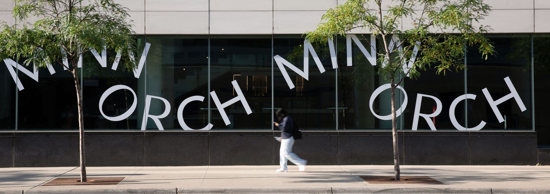 Street view of a modern building with large glass windows displaying the text "MINN ORCH" in a wavy, staggered arrangement, with two small trees on the sidewalk and a blurred person walking by.