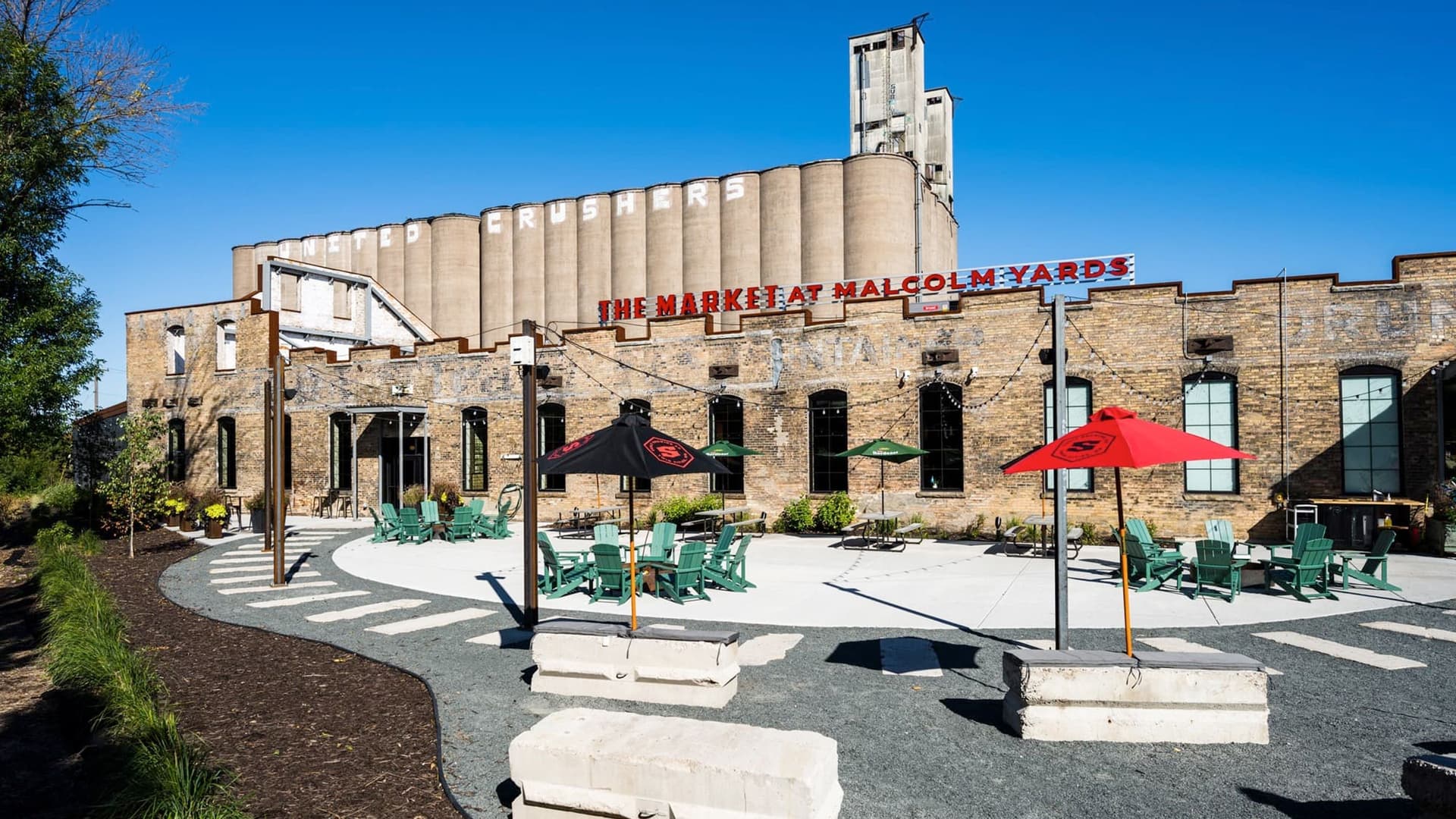 Outdoor seating area with green chairs and umbrellas in front of a brick building with the sign "The Market at Malcolm Yards," and large grain silos labeled "United Crushers" in the background under a clear blue sky.