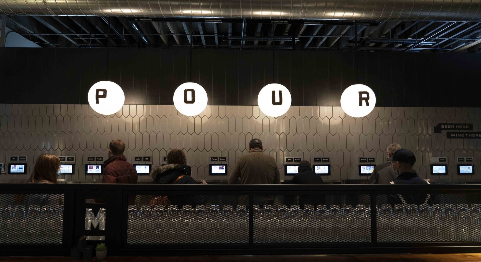 People stand and use digital screens beneath four hanging, illuminated orbs spelling "POUR" in a modern self-serve beverage bar with tiles on the wall.