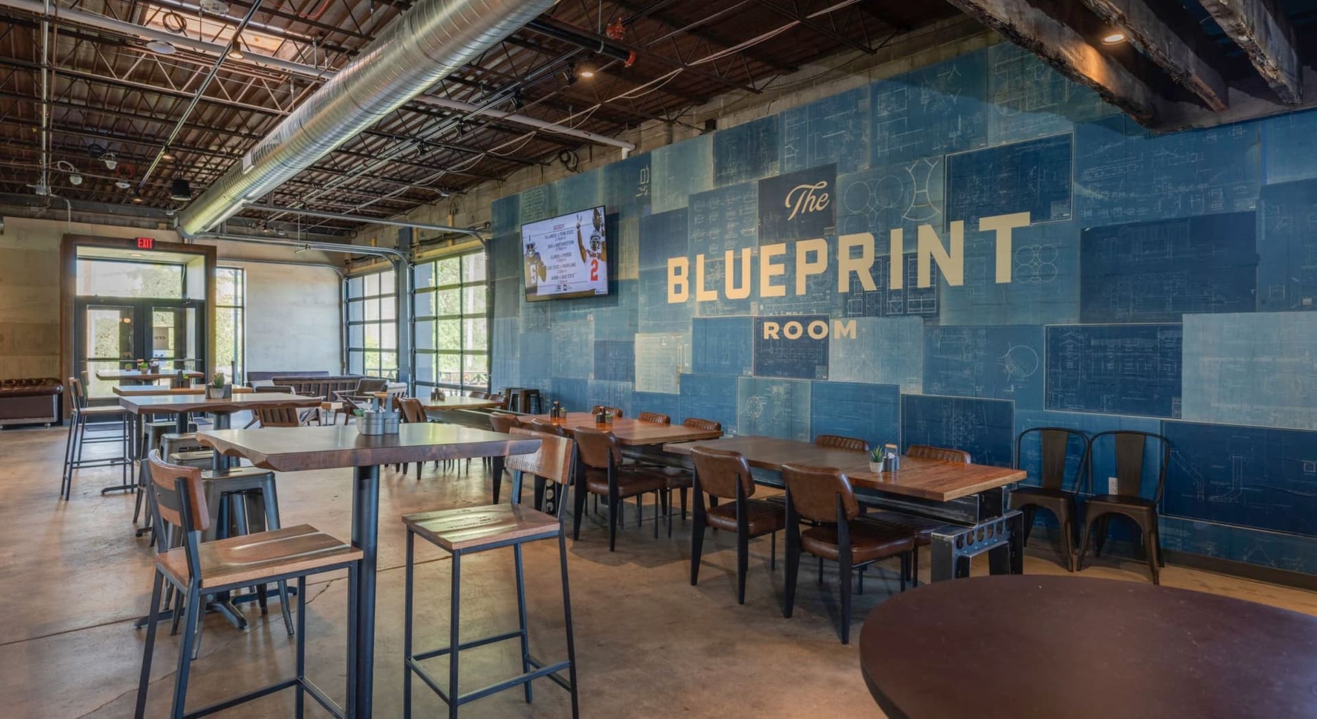 Interior view of a modern dining area called "The Blueprint Room" with wooden tables and chairs, a large blue wall with blueprint designs and the room's name, exposed ceiling with metal ductwork, garage-style windows, and a flat screen TV mounted on the wall.