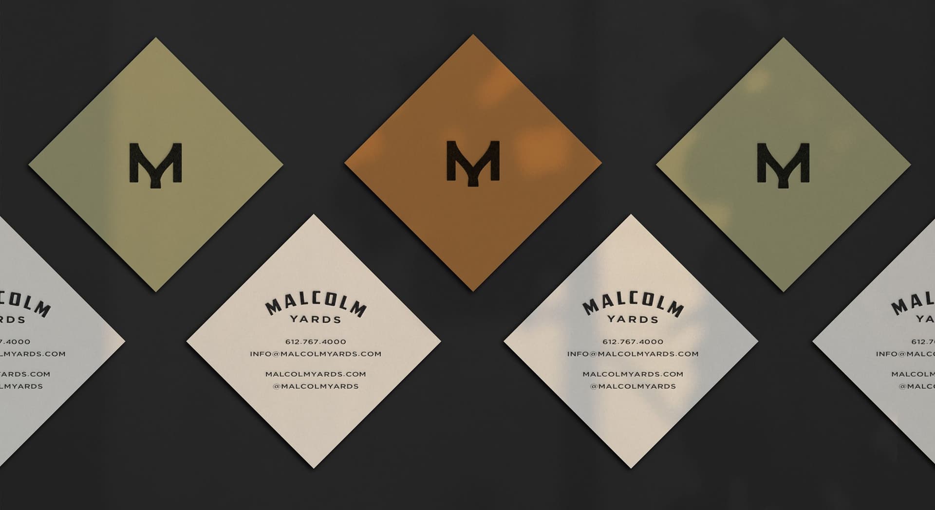 Diamond-shaped business cards arranged in a row on a dark surface. Three cards show the initials "MY" in black on earthy green and burnt orange backgrounds, alternating with three cream-colored cards displaying contact information for Malcolm Yards, including a phone number, email, website, and social media handle.