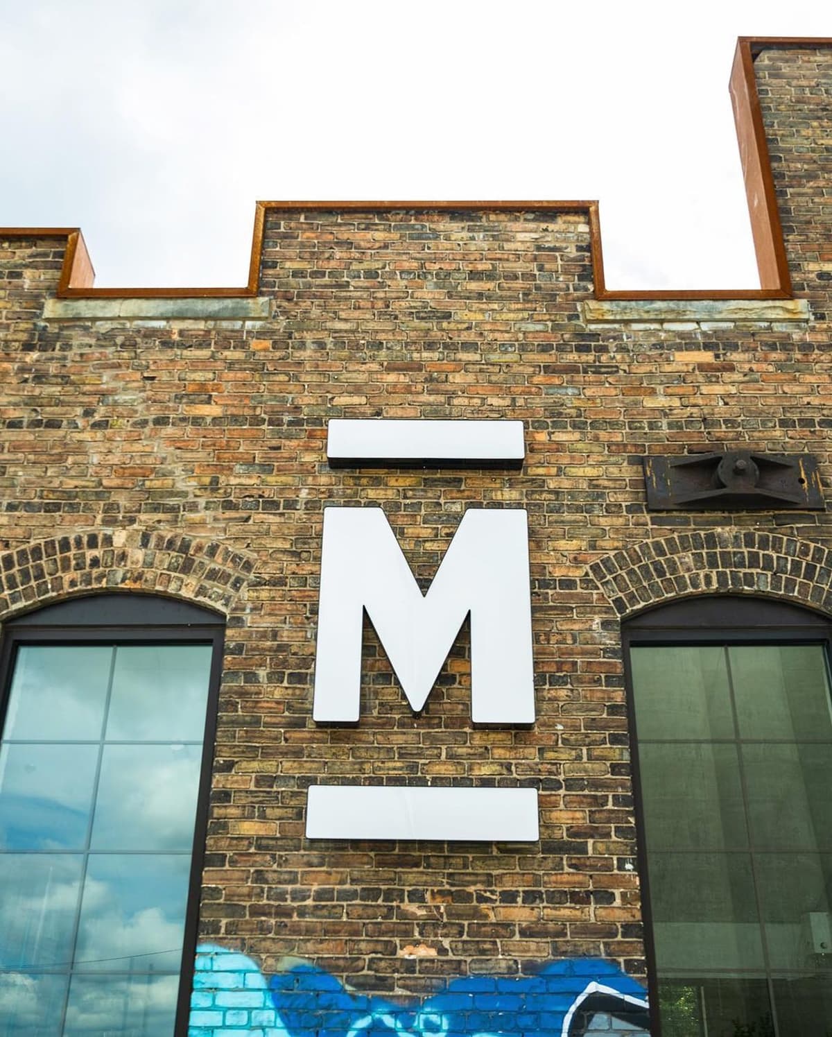 White letter "M" with a horizontal bar above and below it mounted on a brick wall between two arched windows.