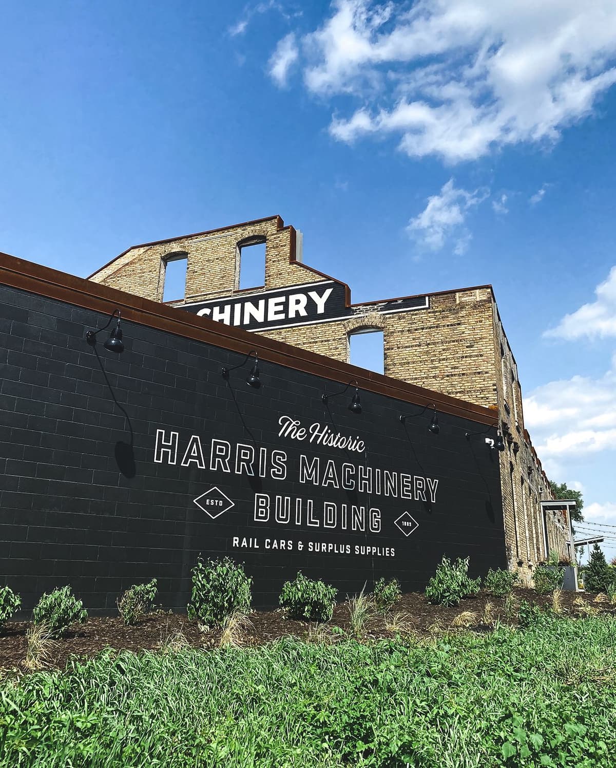 Exterior view of the historic Harris Machinery Building with a black wall featuring white painted text reading "The Historic Harris Machinery Building, Rail Cars & Surplus Supplies, ESTD 1889." The upper part of the building is made of light-colored brick with open, windowless sections, set against a bright blue sky with scattered white clouds. Green plants and grass are visible in the foreground.