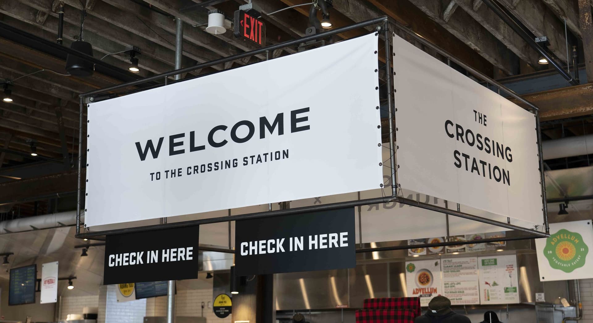 White hanging rectangular banners with black text saying "WELCOME TO THE CROSSING STATION" and "THE CROSSING STATION" in an industrial-style indoor space, with additional smaller black signs below that read "CHECK IN HERE."