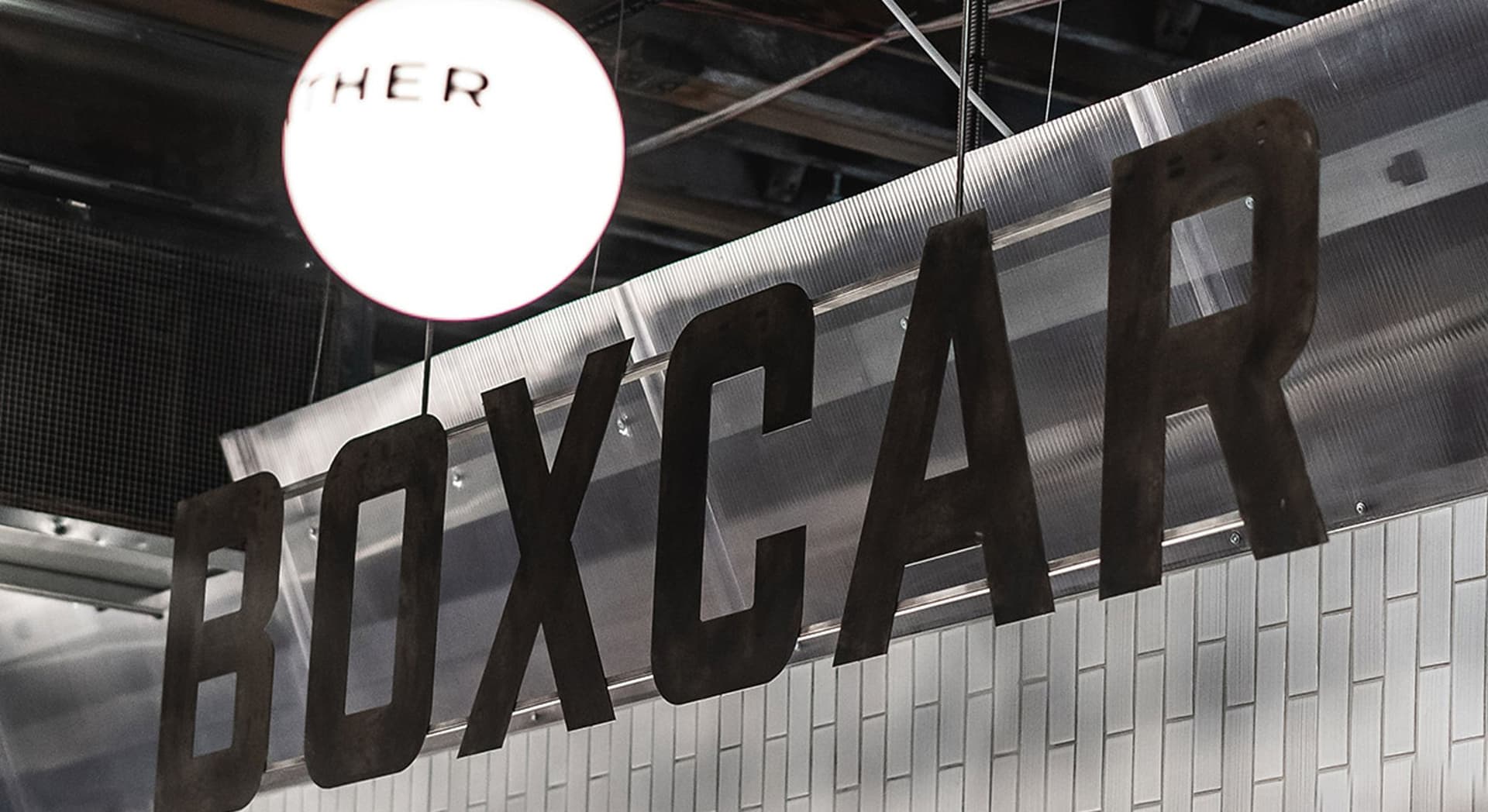 Metal letters spelling "BOXCAR" hanging from the ceiling, with a round light fixture above them and a tiled wall in the background.