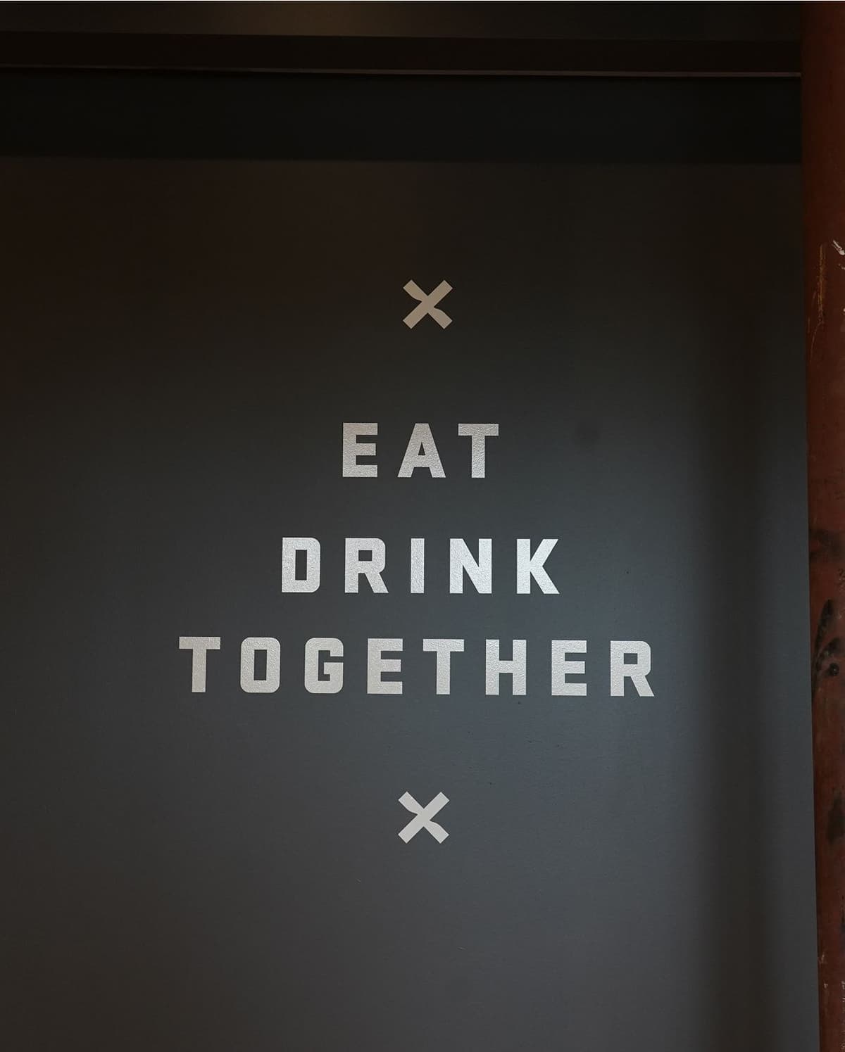 Wall sign with white text on a dark background that reads "EAT DRINK TOGETHER," with white X marks above and below the text.