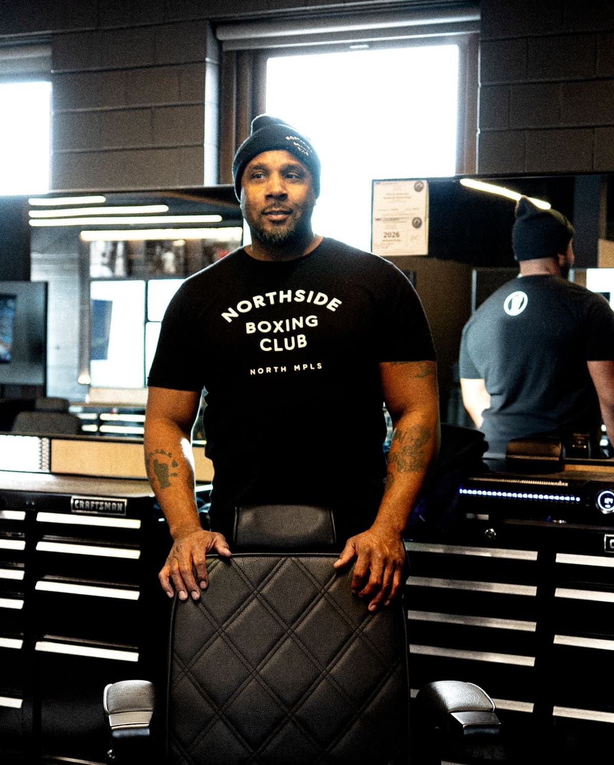 Man wearing a black "Northside Boxing Club" t-shirt and black beanie, standing behind a black leather chair in a gym or workshop setting.