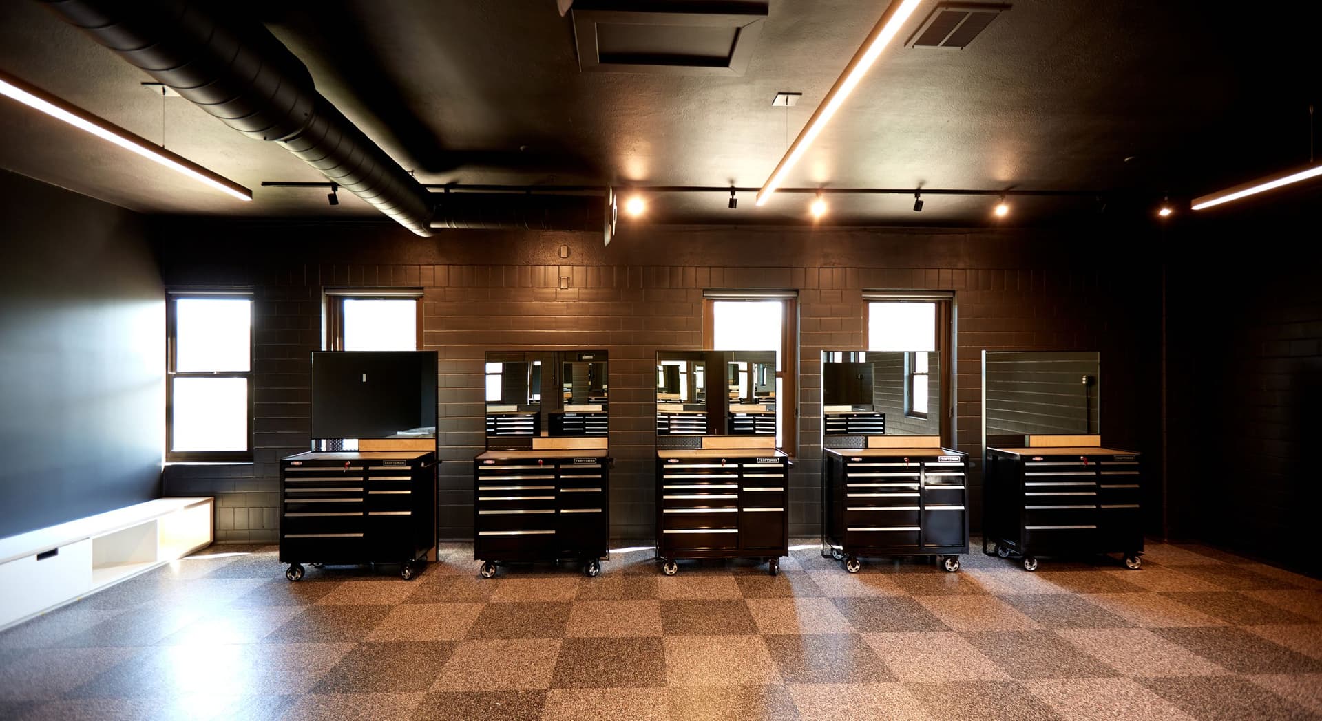 Row of five large black tool cabinets with mirrors on top, positioned in front of four windows on a brick wall inside a well-lit room with a checkered floor and black ceiling.