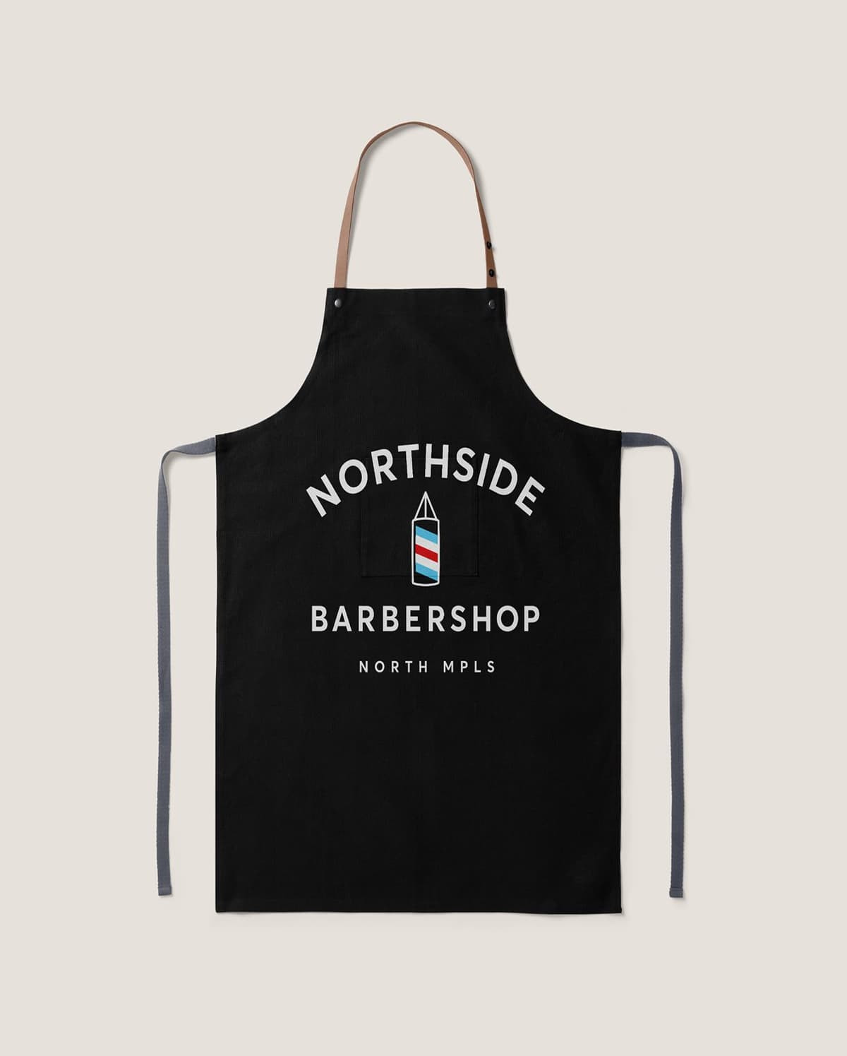 Black apron with tan neck strap and gray waist ties, featuring a graphic of a barber pole and the text "Northside Barbershop North MPLS" printed in white on the front.