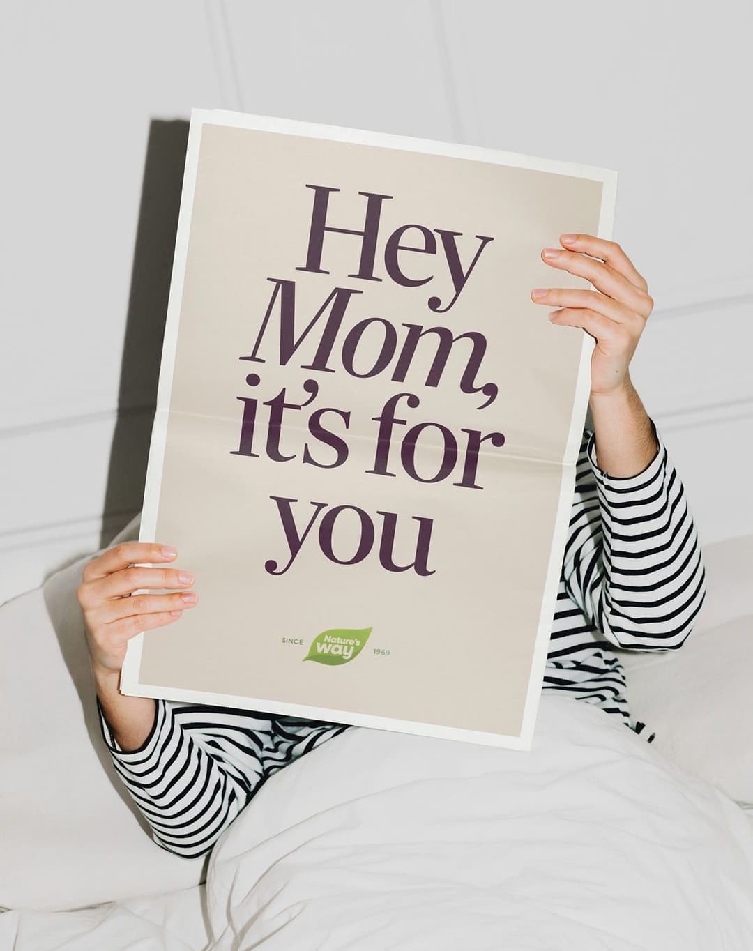 Person in a striped long-sleeve shirt holding a large card that reads "Hey Mom, it's for you" with a Nature's Way logo at the bottom, while sitting in bed under white blankets.