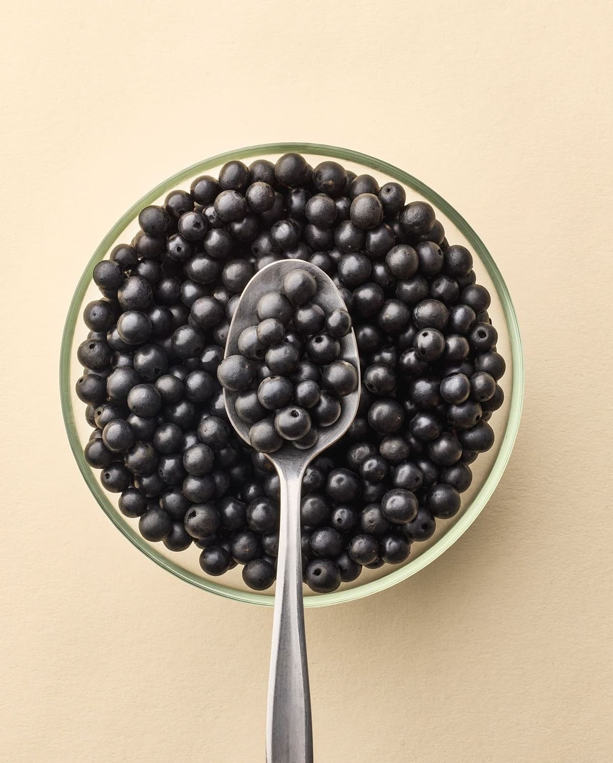 Glass bowl filled with small, round black berries with a spoon holding some of the berries resting on top, against a beige background.