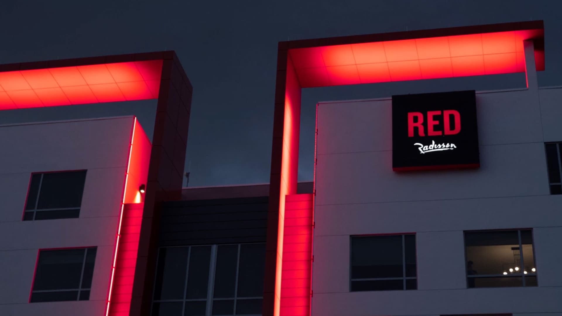 Night view of a modern building with prominent red neon lighting outlining architectural features and a sign that reads "RED Radisson." The facade has several windows, and one window on the right shows a person inside with a chandelier light.