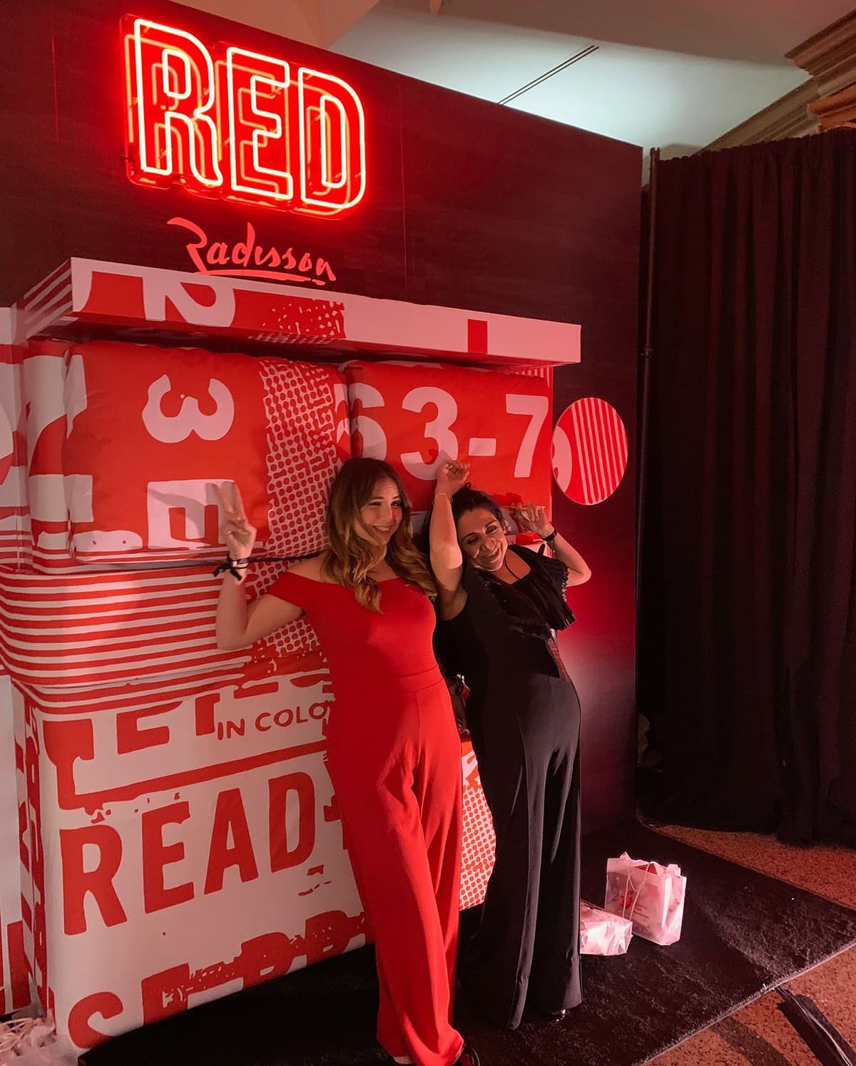 Two women posing with playful peace signs in front of a large red and white sign that reads "RED Radisson," with one woman wearing a red jumpsuit and the other wearing a black dress, standing on a black carpet with gift bags nearby.