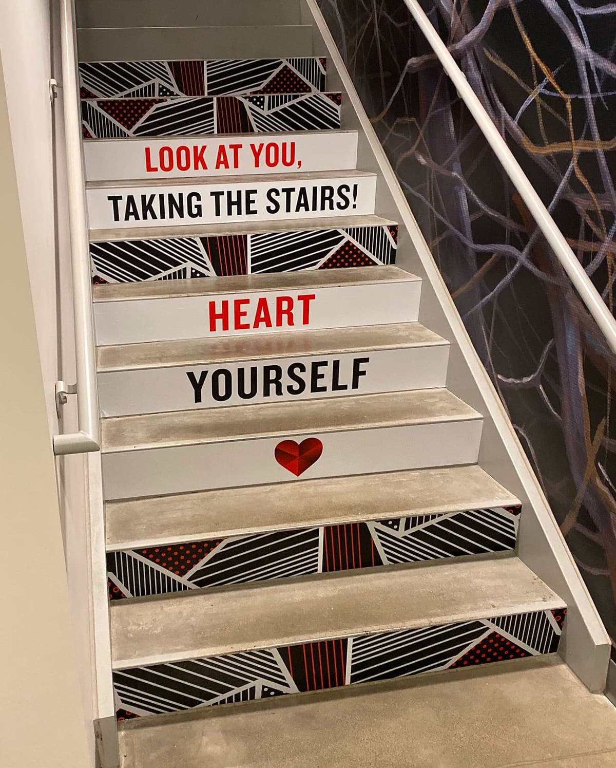 Staircase with motivational text on the risers that reads "LOOK AT YOU, TAKING THE STAIRS! HEART YOURSELF" along with a red heart symbol, featuring a geometric black, white, and red pattern on the sides.