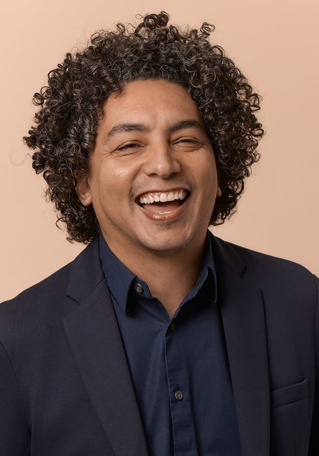 Portrait of a man with curly dark hair wearing a navy blue shirt and dark blazer, smiling broadly against a beige background.