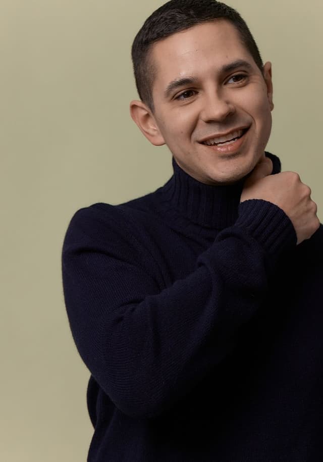 Portrait of a smiling young man with short dark hair wearing a dark navy blue turtleneck sweater against a beige background.