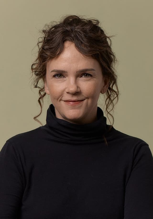 Portrait of a woman with curly brown hair styled up, wearing a black turtleneck sweater, smiling softly against a plain beige background.