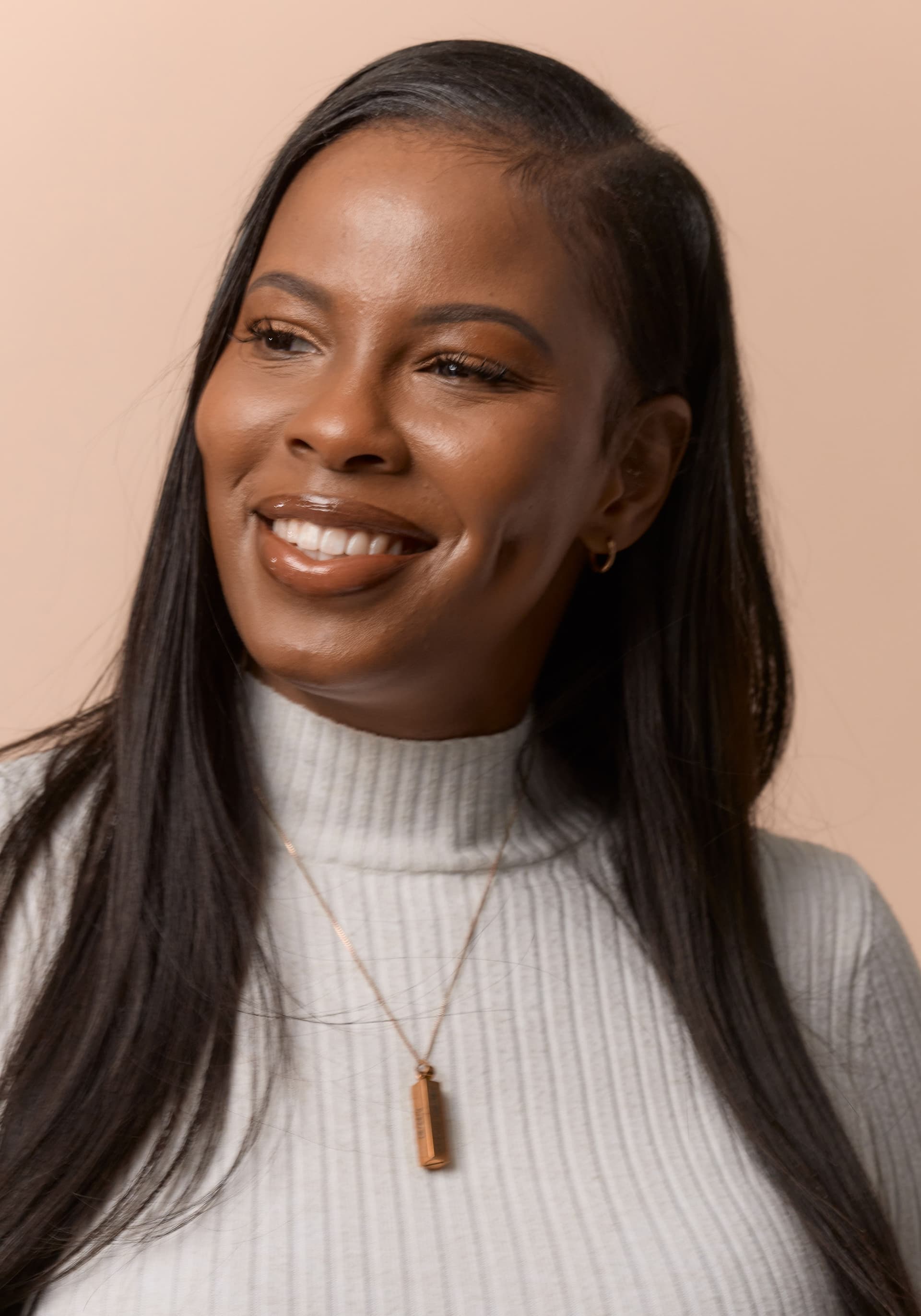 Close-up portrait of a smiling woman with long black hair, wearing a light gray ribbed turtleneck sweater and a thin gold necklace with a rectangular pendant, against a plain beige background.