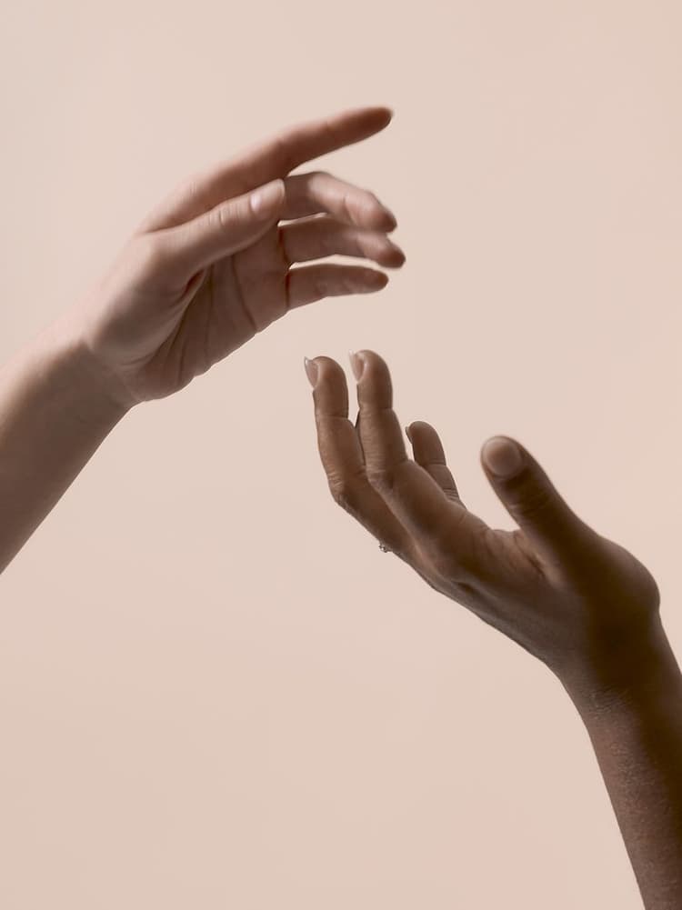 Two hands, one light-skinned and one dark-skinned, reaching towards each other against a neutral beige background.