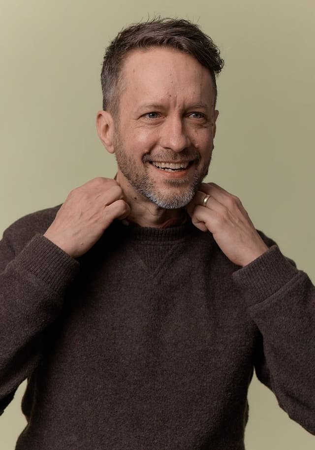 Smiling man with short hair and a beard wearing a dark brown sweater, adjusting his collar against a plain beige background.