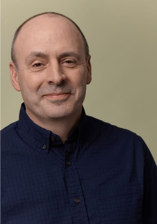 Portrait of a middle-aged man with a receding hairline, wearing a dark blue button-up shirt, smiling softly against a plain beige background.