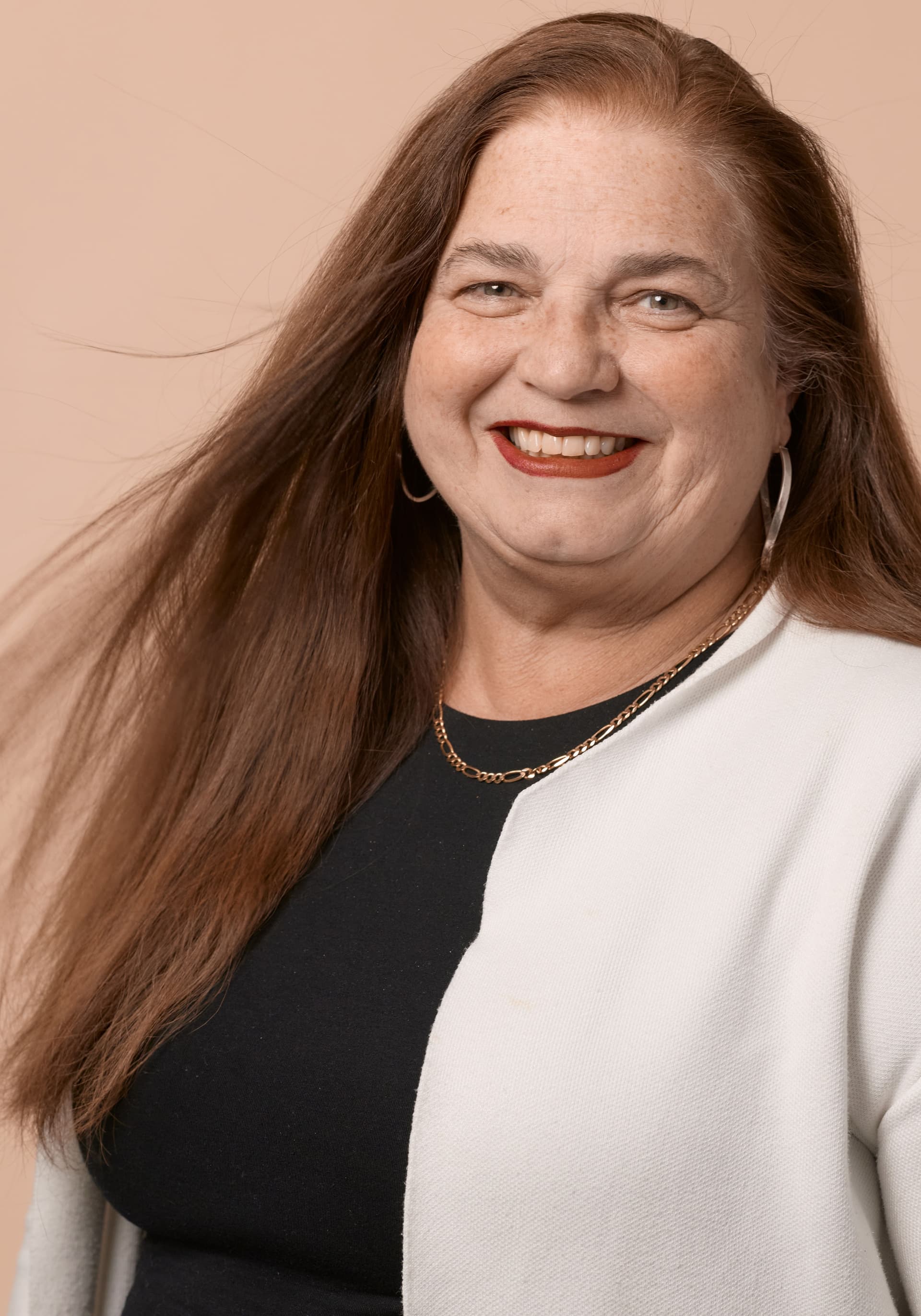 Portrait of a smiling middle-aged woman with long brown hair wearing a black top and white cardigan, gold necklace, and hoop earrings against a beige background.