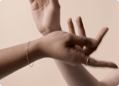 Close-up of two hands with a soft focus, one hand adorned with a delicate gold bracelet and ring, set against a neutral beige background.