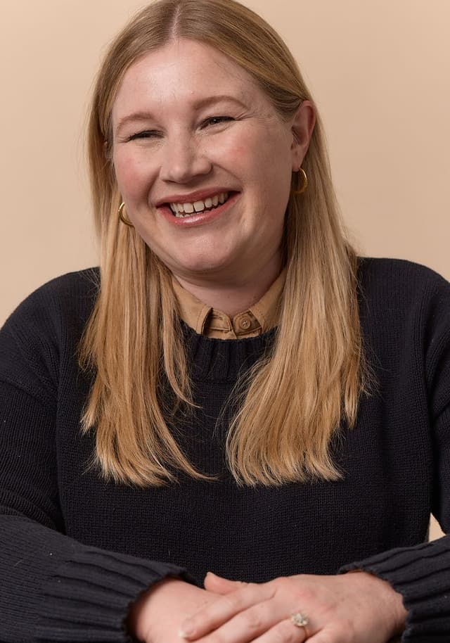 Smiling woman with long blonde hair, wearing a black sweater and brown collared shirt, sitting with hands folded against a beige background.