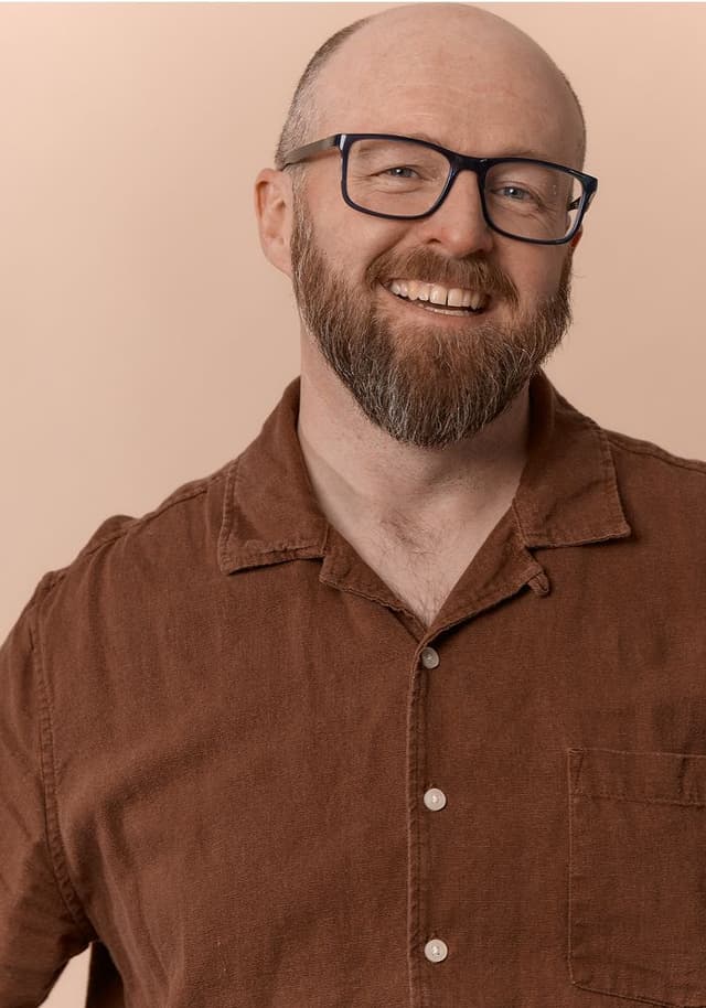 Smiling bald man with a beard and glasses wearing a brown button-up shirt against a beige background.