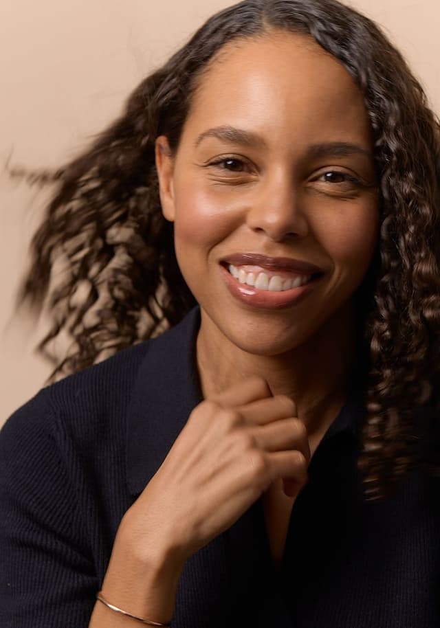 Close-up portrait of a smiling woman with curly hair wearing a dark top, lightly touching her chin, against a plain beige background.