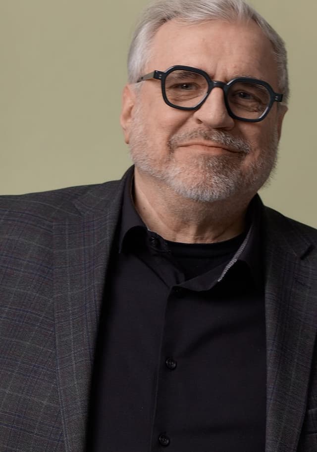 Portrait of an older man with gray hair and a beard, wearing geometric black glasses, a dark plaid blazer, and a black button-up shirt, against a plain beige background.