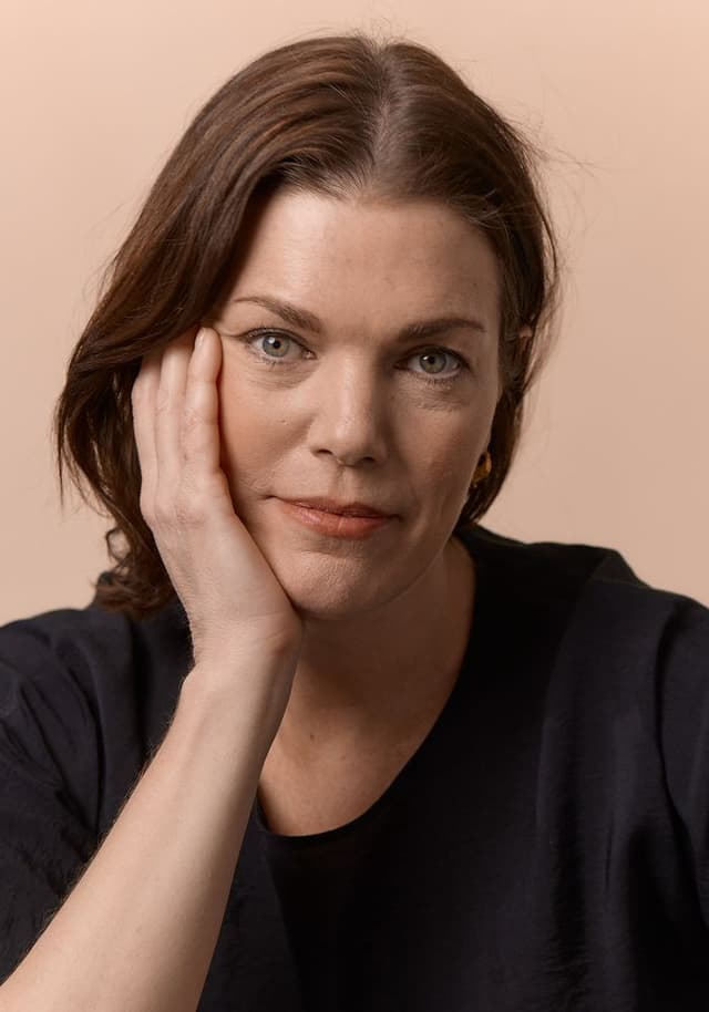 Close-up portrait of a woman with light skin and brown hair resting her face on her right hand, wearing a black top against a beige background.